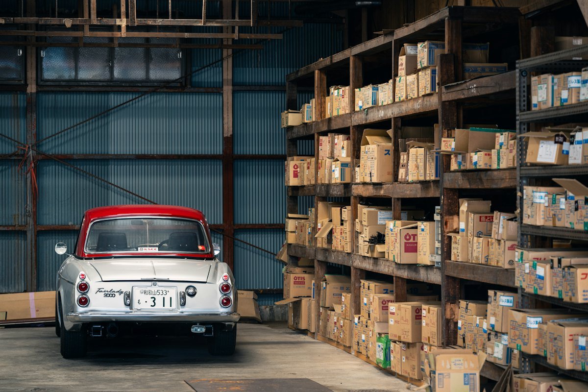 Warehouse racking stacked with boxed parts alongside a parked car.